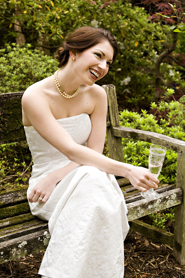 beautiful bride wearing strapless ivory gown and pearls sitting on wooden bench in a garen drinking champagne - photo by North Carolina based wedding photographers Cunningham Photo Artists